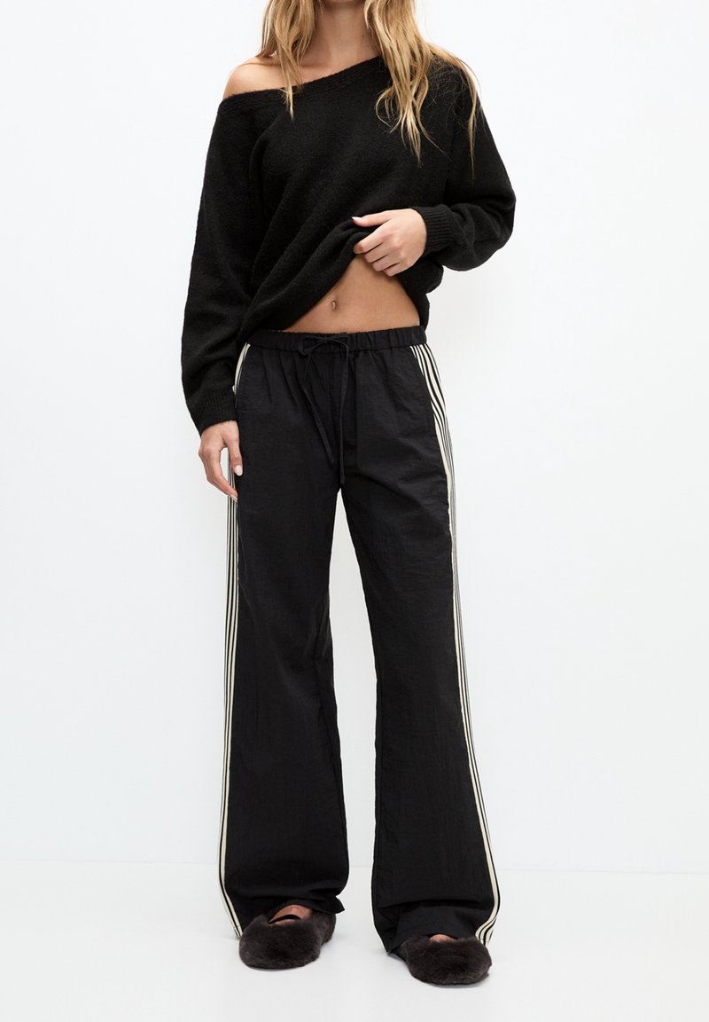 Zwart off-shoulder trui gecombineerd met zwarte wijde broek met witte zijstreep. Model draagt zwarte fluffy pantoffels.