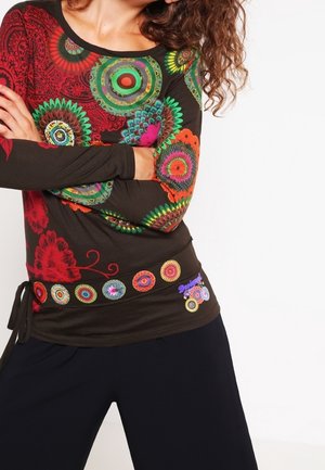 Žena s překříženýma rukama, oblečená v černém tričku s dlouhým rukávem s barevnými kruhovými mandalovými vzory a černých kalhotách.