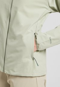 Giacca softshell verde chiaro con zip frontale, tasche laterali, polsini elasticizzati e logo sulla manica. Texture liscia e design moderno.