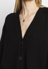 Černý pletený svetr s výstřihem do V, který má na přední části tři knoflíky. Je doplněn zlatým náhrdelníkem s přívěskem ve tvaru slzy.