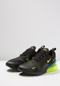 Svarta sportskor med ett andningsbart nätöverdel, neongröna accenter, texturerade detaljer och en synlig luftdämpning i en gradient från blått till gult.