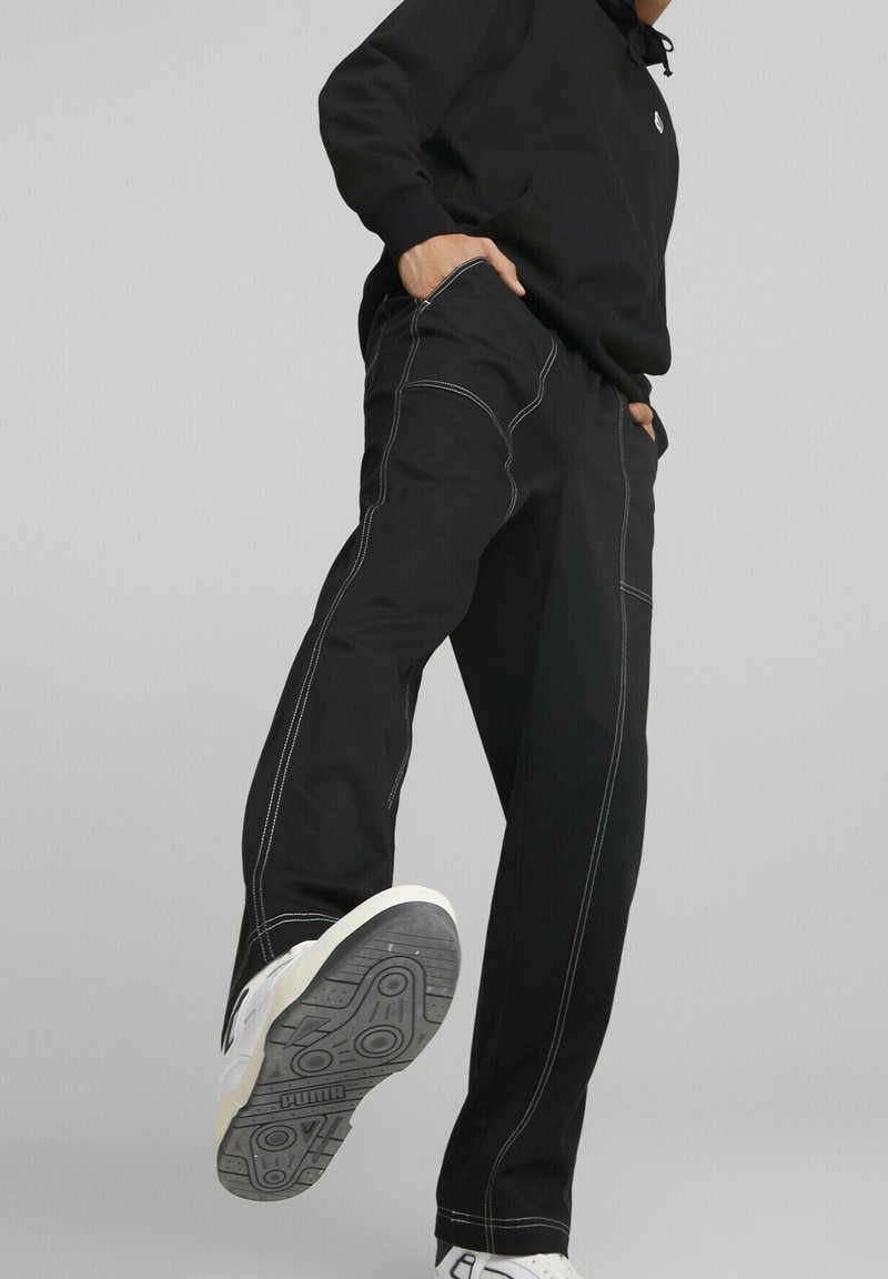 Personne portant un pantalon noir avec des coutures blanches et des baskets blanches Puma, levant une jambe vers l'avant sur un fond gris.