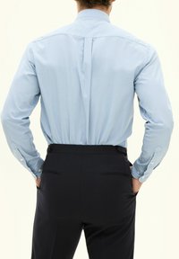 Hellblaues Langarmhemd mit Knopfbündchen und Rückenschlitz, getragen mit dunklen, maßgeschneiderten Hosen mit seitlichen Taschen. Sanfte Textur.