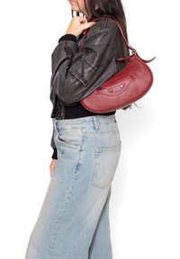 Borsa a spalla rossa in pelle con forma arrotondata, superficie testurizzata e tasca con zip di piccole dimensioni. Abbinata a una giacca bomber nera e jeans azzurri chiari.