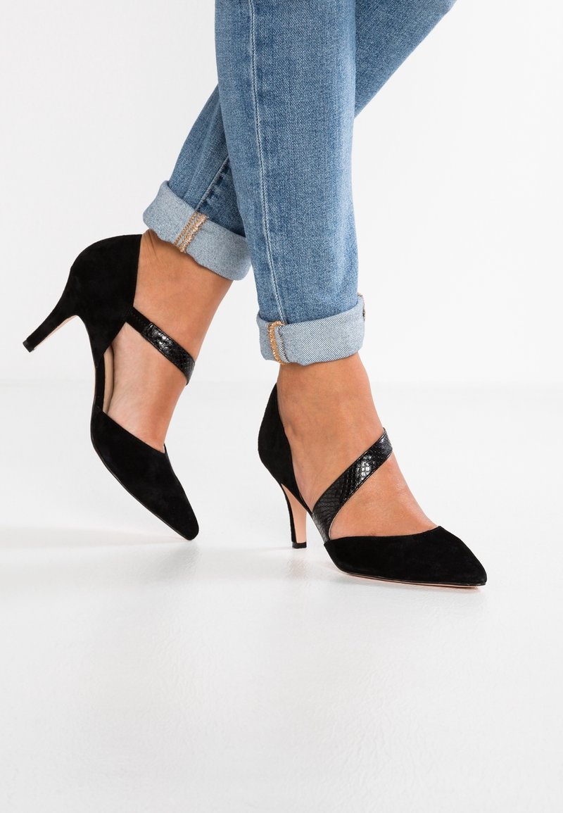 Czarne zamszowe buty na wysokim obcasie z wydłużonym noskiem i paskiem na kostce, charakteryzujące się teksturowanym, błyszczącym wykończeniem. Noszone z podwiniętymi niebieskimi dżinsami.