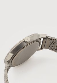 Ασημένιο μεταλλικό ρολόι χειρός που φαίνεται από το πλάι, με κλειστή πίσω θήκη και τοποθετημένη την επιγραφή "ck" στο κουμπί ρύθμισης.