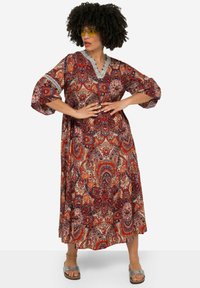 Vestido maxi de patrones rojos y beiges con escote en V, mangas de tres cuartos y detalles de encaje. Lleva sandalias plateadas.
