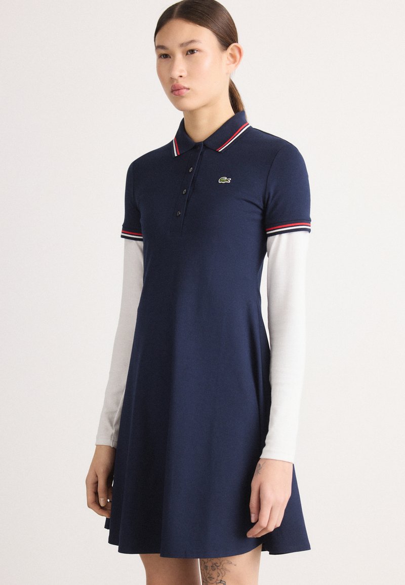 Námořnické polo šaty s červenými a bílými pruhy na límci, krátké rukávy a rozšířený lem; logo Lacoste na hrudi. Hladká bavlněná tkanina.