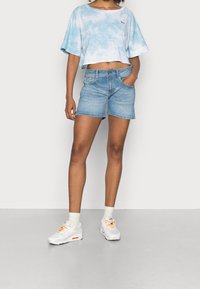 Blue tie-dye crop top with short sleeves, paired with light wash denim shorts. White sneakers with orange accents and light socks.