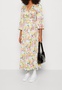 Blommig wrap-klänning med v-ringning, 3/4 ärmar med klockform, färgglada rosor på ljusblått tyg, i kombination med vita sneakers och en svart handväska.