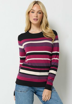 Vrouw met lang blond haar die een gestreepte trui draagt in zwart, paars, roze en crème, gecombineerd met een blauwe spijkerbroek, staande tegen een witte achtergrond.