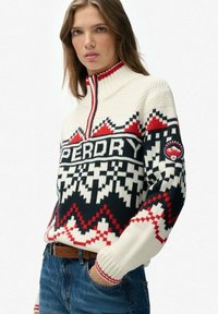 Sweter pulower z dzianiny w kolorze kremowym z czarnymi i czerwonymi geometricznymi wzorami, z wysokim kołnierzem i zapięciem na zamek. Z przodu znajduje się logo "SUPERDRY".
