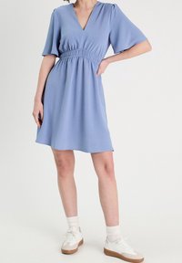 Lyseblå kjole med V-udskæring og elastisk talje, korte flæseærmer, flydende stof, kombineret med hvide sneakers og ankelstrømper.