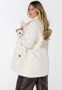 Vrouw met lang blond haar in een witte jas en donkerbruine broek, die zijwaarts poseert tegen een effen witte achtergrond.