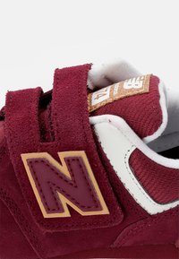 Sneaker in suede bordeaux con dettagli bianchi, fascia in velcro, colletto imbottito e logo dorato. Caratterizzato da materiali testurizzati e cuciture rinforzate.