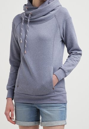 Kvinde iført en grå hættetrøje med snøre og forlommer, kombineret med lyseblå denimshorts med opbrettede ben, stående mod en hvid baggrund.