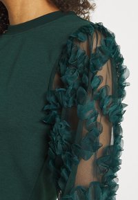 Dark green sweatshirt featuring a solid body and sheer mesh sleeves adorned with layered fabric ruffles in matching shades.