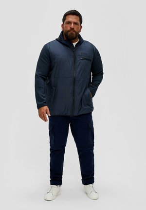 Marineblauen isolierten Jacke mit Reißverschluss, glatter Textur und Seitentaschen. Kombiniert mit marineblauen Cargo-Hosen und weißen Sneakers.