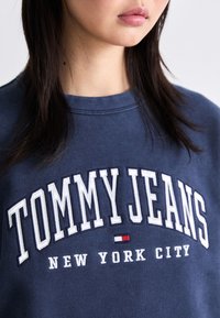 Laivastonsininen huppari, jossa on pyöreä kaula-aukko. Etupuolella on suuri valkoinen teksti, jossa lukee "TOMMY JEANS" ja "NEW YORK CITY" pienillä kirjaimilla.