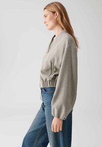 Giacca cropped a spina di pesce grigia con orlo arricciato e maniche voluminose, indossata su jeans blu, con chiusura frontale a zip.