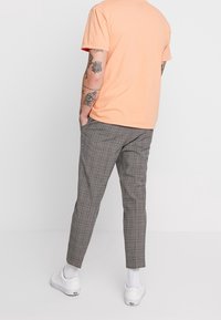 T-shirt pêche à manches courtes associé à un pantalon gris à carreaux. Baskets blanches visibles, avec des tatouages sur le bras visible. Vue arrière.