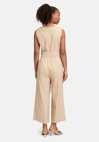 Combinaison en lin beige avec un design à jambes larges, sans manches, taille ceinturée et texture subtile. La vue arrière montre un détail de fermeture à boutons.