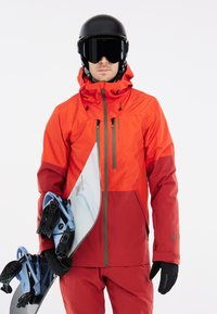 Orangen und rote wasserdichte Jacke mit Kapuze, grauen Reißverschlüssen und mehreren Taschen; er hält ein Snowboard mit blauen Bindungen. Schwarzer Helm und Handschuhe.