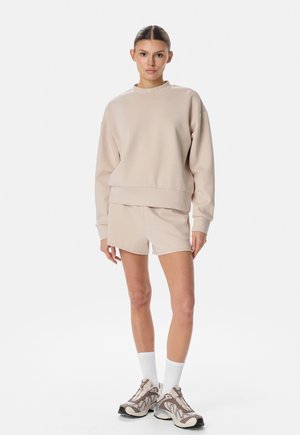 Kobieta stoi, ubrana w beżową bluzę z długim rękawem, pasujące szorty, białe skarpetki za kostkę oraz beżowe sportowe buty na białym tle.