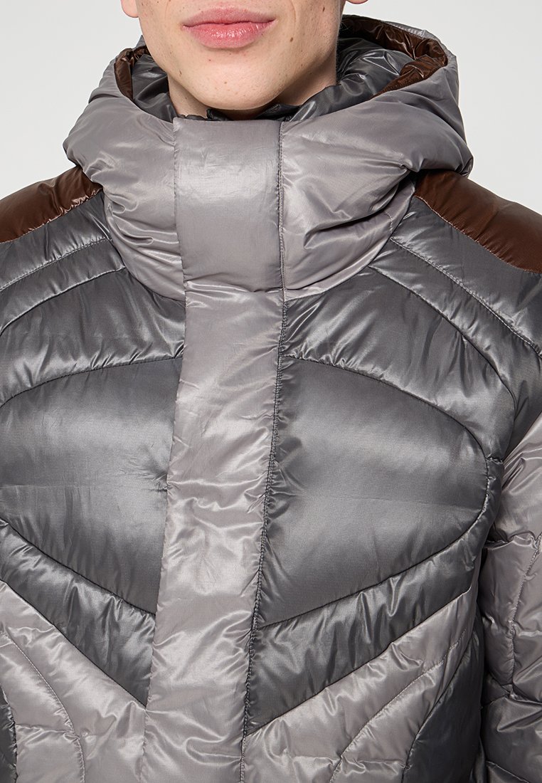 Chaleco acolchado con un alto cuello, que presenta un diseño en forma de flecha en tonos de gris y marrón. Tejido suave y brillante con textura acolchada.