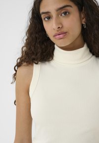 Jonge vrouw met lang krullend haar, gekleed in een witte geribbelde mouwloze coltrui en zilveren oorringen, tegen een effen achtergrond.