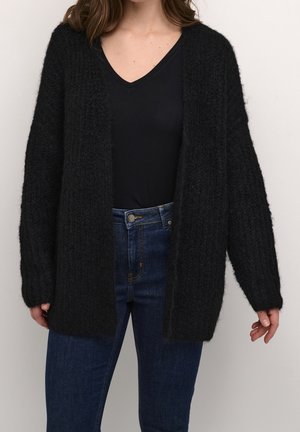 Femme portant un cardigan noir en tissu duveteux sur un haut noir à col en V et un jean taille haute bleu, debout devant un fond uni.