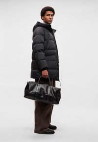 Casaco acolchoado preto com capuz, combinado com uma mala de viagem em couro preto trançado, com duas alças e acabamento liso. Calças e sapatos castanhos.