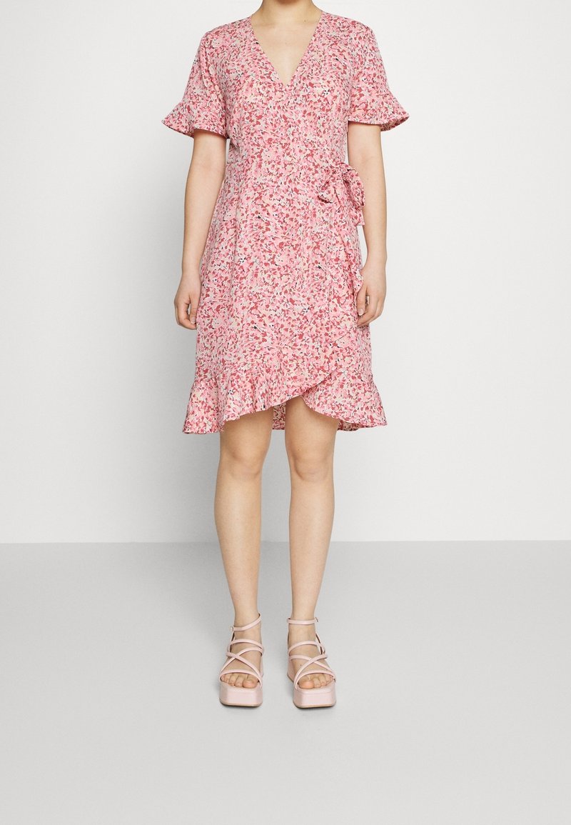 Femme portant une robe portefeuille fleurie rose longueur genou avec des manches courtes volantées et un ourlet à volants, associée à des sandales compensées roses.
