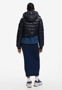 Giacca corta imbottita nera con cappuccio, texture lucida e cuciture orizzontali, indossata sopra una giacca di jeans, abbinata a una gonna a coste blu navy.