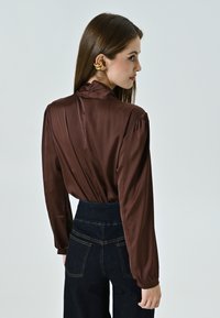 Blusa in satin marrone con colletto alto, maniche a sbuffo e retro arricciato, abbinata a jeans in denim scuro a vita alta con tasche posteriori.