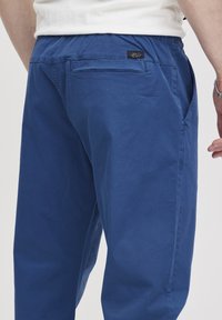 Niebieskie bawełniane spodnie o luźnym kroju, z tylną kieszenią i elastycznym pasem. Materiał wydaje się gładki, o minimalnej fakturze.