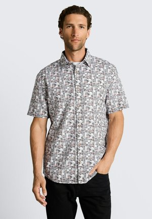 Mann trägt ein kurzärmliges Hemd mit schwarzem und rotem abstraktem Blattmuster, kombiniert mit einer schwarzen Hose, stehend vor hellem Hintergrund.