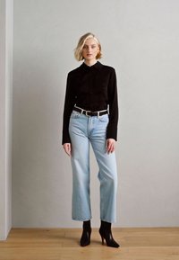 Chemise noire à manches longues avec des boutons, jean taille haute bleu clair, ceinture blanche et bottines noires. Le mannequin se tient devant un fond neutre.