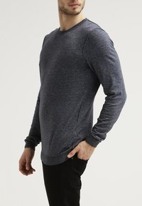 Mann in einem dunkelgrauen Langarmpullover mit gerippten Bündchen und Saum, kombiniert mit schwarzen Hosen, steht vor einem schlichten Hintergrund.