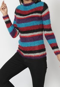 Pull multicolore avec des rayures horizontales bleu, rouge, violet, noir et gris. Fabriqué à partir d'un matériau moelleux et texturé, avec un col montant.