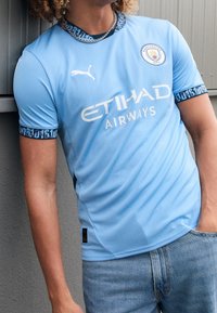 Vaaleansininen lyhythihainen urheilupaita, jossa mustia kuvioituja yksityiskohtia kauluksessa ja hihoissa. "ETIHAD AIRWAYS" painettuna valkoisella.