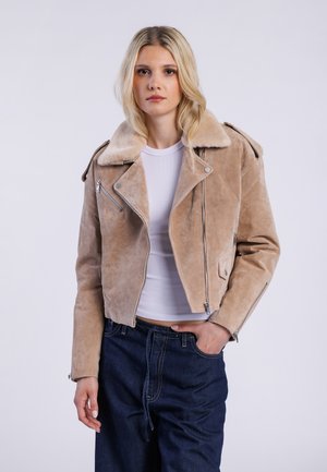 Veste de biker en suède beige avec un col en fausse fourrure doux, fermetures éclair, et design ajusté, associée à un haut blanc et un jean bleu foncé.