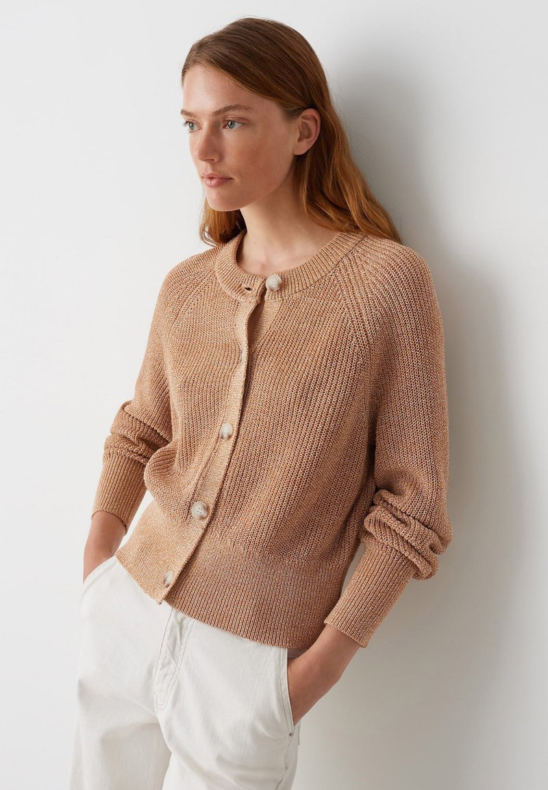 Hellbrauner Strickcardigan mit rundem Ausschnitt, Knopfverschluss, gerippten Bündchen und strukturierter Oberfläche. Kombiniert mit weißen Hosen.
