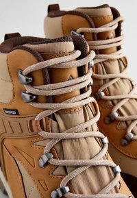 Brązowe i beżowe buty trekkingowe mają wodoodporną cholewkę, tekstylny kołnierz oraz sznurowanie z metalowymi haczykami i teksturowanymi akcentami.