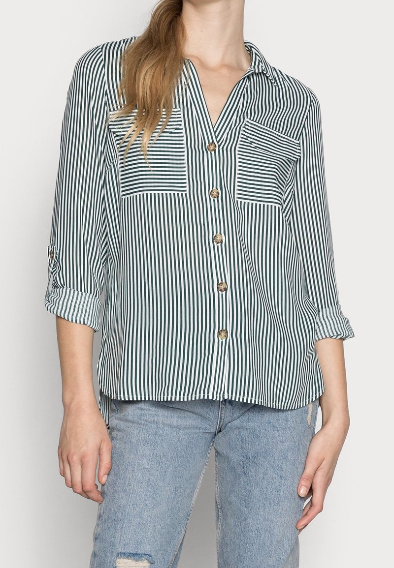 Chemise boutonnée à rayures vertes et blanches, dotée de deux poches poitrine, d'un col en V et de manches retroussées avec des boutons.