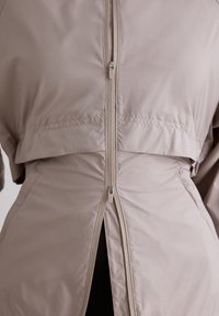 Veste beige à fermeture éclair bidirectionnelle avec fermetures visibles, partiellement ouverte en bas laissant entrevoir une couche intérieure noire.