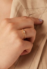 Goldring mit einem hexagonalen Stein und einer leichten Textur. Der Ringboden weist ein zartes Kettenmuster auf, das vor einem neutralen Stoffhintergrund gesetzt ist.