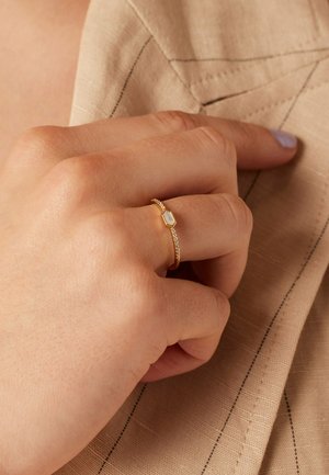 Gouden ring met een hexagonale steen met een lichte textuur. De band heeft een delicaat kettingpatroon, tegen een neutrale stofachtergrond.