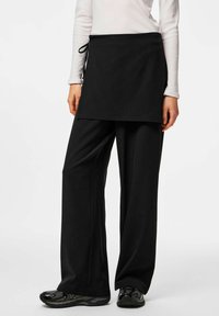 Tablier noir avec une ceinture à nouer à la taille, accompagné d'un pantalon noir ample. La tenue présente une texture lisse et un design minimaliste.