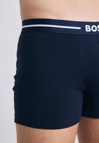 Navy blue cotton boxer shorts with a fitted design, featuring a contrasting white logo band at the waist and smooth texture.